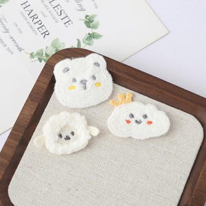 Puede incluir: Tres parches bordados: un oso blanco con detalles amarillos, una nube blanca con una corona amarilla y una oveja blanca. Los parches están sobre una tela beige dentro de una bandeja de madera. Una invitación de boda está en el fondo.