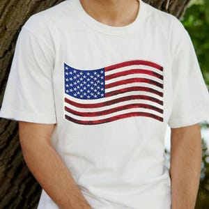 May include: White t-shirt featuring a sequined American flag design. The flag has red and white stripes and a blue field with white stars. The shirt is short-sleeved and appears to be made of cotton.