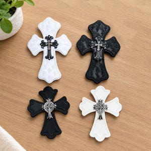 May include: Four cross-shaped patches arranged in a square. Two are white with black metal cross emblems, and two are black with silver metal cross emblems. The patches feature embossed patterns and scalloped edges, showcasing detailed metal hardware.