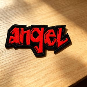 以下が含まれることがあります： 鮮やかな赤色の糸で刺繍されたangelという文字の布製ワッペンで、太い黒色の縁取りが施されています。文字は太くブロック体のようなフォントで、はっきりとした輪郭線が描かれており、コントラストの強いデザインに仕上がっています。