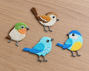 Könnte beinhalten: Vier gestickte Vogelaufnäher mit detaillierten Nähten. Ein Vogel ist braun-weiß, einer grün-orange, einer hellblau und einer leuchtend blau-gelb. Jeder Aufnäher hat einen kleinen schwarzen Schnabel, ein schwarzes Auge und zwei dünne Beine.