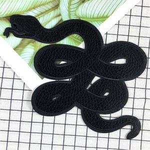 May include: A black snake-shaped embroidered patch. The patch features a detailed design of a coiled snake with a textured surface. The patch is likely for decorative purposes, such as adding to clothing or accessories.