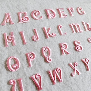 Könnte beinhalten: Rosa gestickte Buchstaben des Alphabets, A bis Y, auf hellem Stoff angeordnet. Jeder Buchstabe hat ein dekoratives Wirbeldesign. Die Buchstaben sind gleichmäßig angeordnet und scheinen zum Aufbügeln oder Aufnähen zu sein.