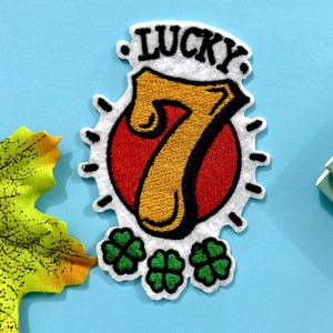 Może przedstawiać: Biała, haftowana naszywka z napisem "LUCKY" nad złotą cyfrą 7, na czerwonym okręgu. Naszywka zawiera trzy zielone koniczyny i czarny kontur. Naszywka znajduje się na jasnoniebieskim tle.