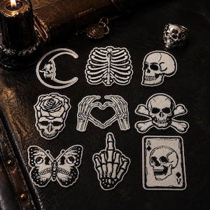 May include: A collection of eight embroidered patches featuring black and white designs including a crescent moon with a skull, a rib cage, a skull, a rose with a skull, skeleton hands forming a heart, a skull and crossbones, a butterfly, and an ace card.