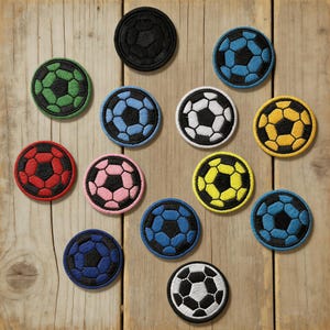 Può includere: Una collezione di toppe ricamate a forma di pallone da calcio in vari colori, tra cui nero, verde, blu, bianco, giallo, rosa e rosso. Le toppe sono rotonde con un design dettagliato del pallone da calcio e sono disposte su una superficie di legno.