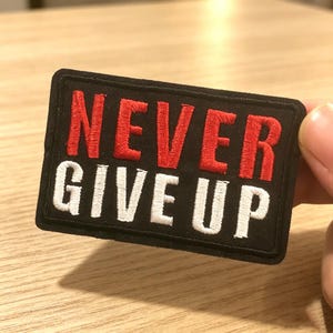 May include: A rectangular black embroidered patch featuring the text NEVER in bold red capital letters on the top line and the text GIVE UP in bold white capital letters on the bottom line, with a black stitched border surrounding the entire design.