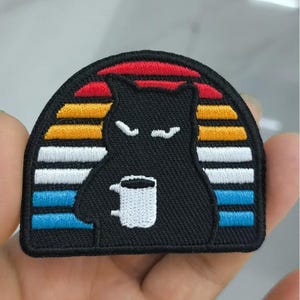 May include: Embroidered patch showing a black cat with a white mug, set against a sunset design with red, orange, yellow, white, and blue stripes. The cat has a grumpy expression. The patch has a black, stitched border.