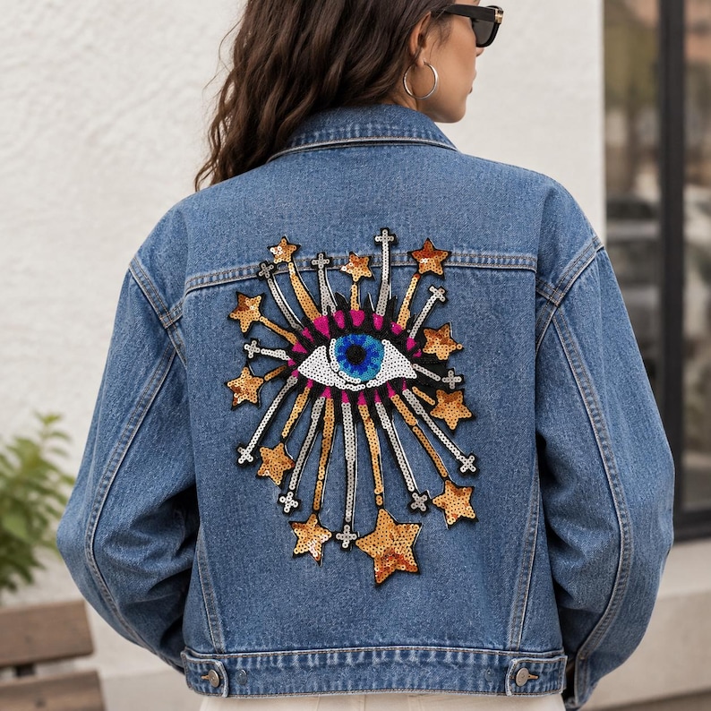 K&ouml;nnte beinhalten: Eine blaue Jeansjacke mit einem gro&szlig;en, bunten Paillettenaufn&auml;her auf der R&uuml;ckseite. Der Aufn&auml;her zeigt ein zentrales Auge mit blauer Iris, umgeben von strahlenf&ouml;rmigen Linien und goldenen Sternen, die aus schimmernden Pailletten bestehen.