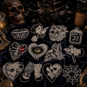 May include: A collection of twelve embroidered patches featuring gothic and occult imagery, including skulls, anatomical hearts, eyes, hands, and spiderwebs. The designs are rendered in black and white thread with occasional red accents on a dark blue velvet fabric.