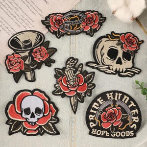 以下が含まれることがあります： バラ、スカル、そして「HOPE GOODS」と「PRIDE HUNTERS」の文字が刺繍されたパッチのコレクション。パッチは黒、赤、白で、詳細なステッチが施されています。デザインには、バラのあるスカル、バラを通して短剣を持つ手、バラのあるメガホンが含まれています。