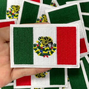 Könnte beinhalten: Gestickter Aufnäher mit der mexikanischen Flagge. Der rechteckige Aufnäher hat einen weißen Mittelstreifen, einen grünen vertikalen Streifen und einen roten vertikalen Streifen. In der Mitte befindet sich das mexikanische Wappen. Die Aufnäher sind weiß mit grünen, roten, gelben und schwarzen Details.