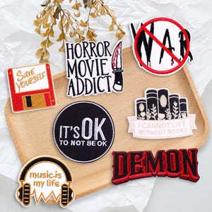 May include: A selection of embroidered patches with diverse designs and text. The patches feature phrases such as "Horror Movie Addict," "It's OK to Not Be OK," and "Music is My Life." Other patches depict a knife, a crossed-out "WAR" symbol, and a floppy disk.