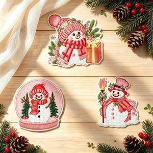 May include: Three embroidered patches featuring snowmen with pink and red accents. One snowman wears a beanie and holds a gift, another is inside a circular snow globe, and the third wears a top hat while holding a lit torch with greenery.