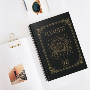 Puede incluir: Un cuaderno de espiral negro con un diseño dorado del signo zodiacal Cáncer en la portada. La portada muestra la palabra CANCER en letras elegantes, una ilustración detallada de un cangrejo, constelaciones y un marco decorativo.