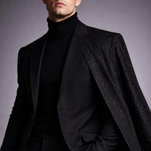 Puede incluir: Un traje negro con un cuello alto negro a juego. La chaqueta del traje presenta un dise&ntilde;o tipo capa con un patr&oacute;n brillante en los hombros. Se ve un reloj plateado.