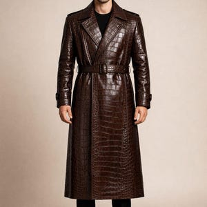 May include: A full-length, brown leather trench coat with a crocodile skin texture. The coat features a belted waist, notched lapels, and buttoned cuffs. The person is wearing a black shirt and black pants.