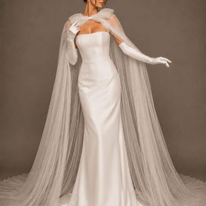 Op de afbeelding: Een witte strapless trouwjurk met een vloeiende sleep. De bruid draagt een doorschijnende, gestippelde cape en lange witte handschoenen. De cape is met een strik aan de schouders bevestigd. De achtergrond is effen en neutraal.