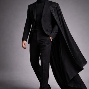Puede incluir: Un traje negro con un cuello alto a juego y una larga capa fluida con un patr&oacute;n brillante. El traje incluye una americana y pantalones a medida. La capa cae dram&aacute;ticamente, agregando un toque de elegancia al conjunto.