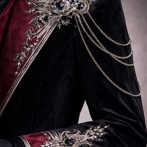 May include: Black velvet jacket with burgundy accents and ornate silver embellishments. Features intricate floral and jewel details on the shoulders and cuffs, with delicate chain accents. A luxurious, gothic-inspired garment.