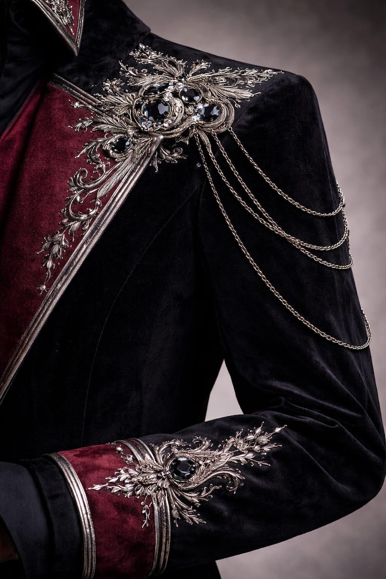 May include: Black velvet jacket with burgundy accents and ornate silver embellishments. Features intricate floral and jewel details on the shoulders and cuffs, with delicate chain accents. A luxurious, gothic-inspired garment.