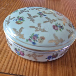 Könnte beinhalten: Eine ovale Schmuckdose aus Porzellan mit einer hellen mintgrünen Grundfarbe. Der Deckel und die Seiten sind mit einem wiederkehrenden Muster aus kleinen, bunten Blumensträußen in Rosa, Blau und Gelb sowie zarten, goldfarbenen Blattmotiven verziert.
