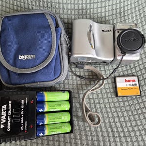 May include: A silver Casio digital camera with a lens cap, a blue Bigben carrying case, a black Varta compact battery charger containing four green rechargeable batteries, and a Hama 128 MB CompactFlash memory card with a grey wrist strap.