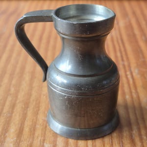 Peut inclure: Un petit pichet gris métallique avec une finition mate et lisse. Le récipient présente un corps arrondi, un col étroit, un bord évasé et une poignée incurvée unique fixée sur le côté. La surface montre des signes d'usure et de légères rayures.