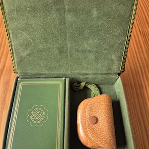 Peut inclure: Boîte verte en velours ouverte, contenant un étui à cartes vert bordé d'or et une petite pochette en cuir marron clair. L'étui à cartes porte un emblème doré. La pochette a un bouton et une sangle verte.