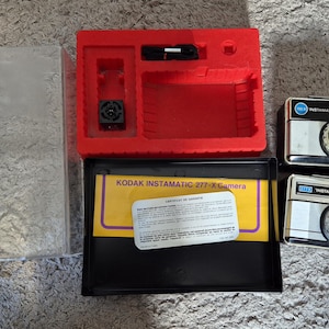 May include: Two Kodak Instamatic 277-X cameras with silver and black bodies, alongside their original packaging. The packaging includes a red plastic insert, a clear plastic cover, and a black box with a yellow label.