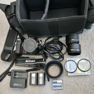 Op de afbeelding: Een zwarte Nikon camerabody met lensdop, een zwarte cameratas, twee Nikon lenzen, een acculader, twee accu's, een zonnekap, twee ronde filters, een 2GB CompactFlash geheugenkaart, een zwarte camerariem en een zwarte stroomkabel.
