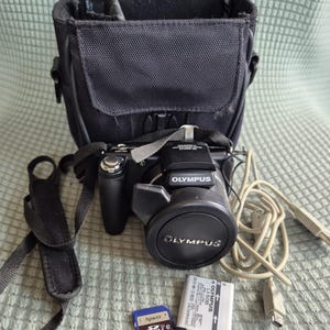 Könnte beinhalten: Eine schwarze Olympus Digitalkamera mit Objektivdeckel, schwarzer Kameratasche, schwarzem Trageriemen, hellgrauem USB-Kabel, silbernem Olympus LI-50B Akku und einer blauen Apacer 32GB SDHC-Speicherkarte. Der Markenname Olympus ist aufgedruckt.