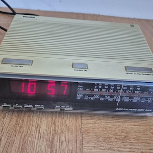 Peut inclure: Un radio-réveil électronique Philips beige avec un affichage numérique indiquant les chiffres rouges 10 57. L'appareil dispose d'un tuner radio horizontal et de trois boutons de commande sur le dessus marqués Alarm Off, Slumber On et Repeat Alarm.