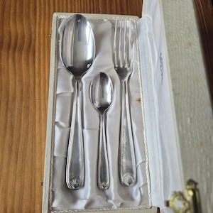 Può includere: Un set di posate color argento, tra cui un cucchiaio, un cucchiaino e una forchetta, presentato in una scatola color crema con fodera in raso. I manici delle posate hanno un motivo a conchiglia.