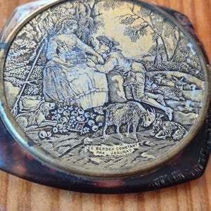 Puede incluir: Un objeto decorativo redondo y vintage con un grabado detallado de una escena pastoral. La imagen presenta figuras, ovejas y árboles, con el texto "LE BERGER CONSTANT PAR JEAURAT" alrededor. Tiene un borde oscuro y estampado.