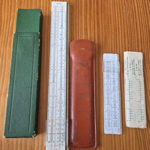 Peut inclure: Ensemble de règles à calcul et accessoires vintage. Comprend un étui vert, une règle à calcul avec des marquages blancs et rouges, un étui en cuir marron avec le numéro 615, une règle à calcul plus petite et un papier avec des calculs.