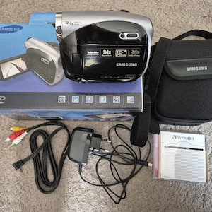 May include: A silver and black Samsung DVD digital camcorder with 34x optical zoom, featuring a Schneider Kreuznach lens. Includes a blue Samsung product box, a black carrying case, a power adapter, AV cables, and a Verbatim DVD disc in a case.