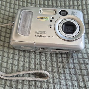 Peut inclure: Un appareil photo numérique argenté Kodak EasyShare CX6330 avec un objectif de 3,1 mégapixels, un flash intégré et une dragonne gris clair. À côté se trouve une carte mémoire SD Panasonic 16 Mo bleue. Le boîtier porte le logo Kodak et du texte.