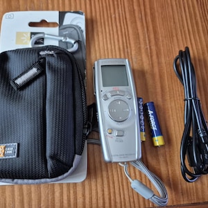 Könnte beinhalten: Ein silberner Olympus Digital-Sprachrekorder mit grauer Handschlaufe, zwei A-Force AAA-Batterien, einem schwarzen USB-Kabel und einer schwarzen Case Logic Reißverschlusstasche mit silbernem Karabiner in der Originalverpackung.