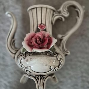 Peut inclure: Une crémière décorative en céramique avec une rose rose en accent. La crémière est blanc cassé avec des rayures verticales et des détails ornés. Elle comporte une poignée incurvée et un bec, et repose sur trois petits pieds.