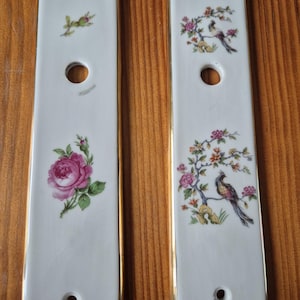 Może przedstawiać: Dwie prostokątne płytki drzwiowe z białej porcelany ze złotymi krawędziami. Lewa płytka przedstawia różową różę i małą zieloną gałązkę. Prawa płytka ukazuje kolorowego ptaka siedzącego na kwitnącej gałęzi. Obie płytki mają trzy okrągłe otwory.