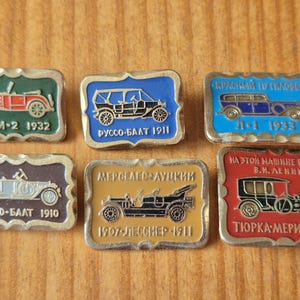 Può includere: Una collezione di sei spille metalliche rettangolari con illustrazioni di auto d'epoca in vari colori tra cui verde, blu, marrone e rosso. Ogni spilla mostra un'automobile classica con testo in cirillico e date come 1932, 1911, 1933, 1910 e 1908.