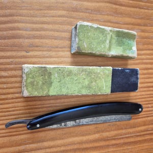 Può includere: Un rasoio a mano libera con manico nero e lama metallica, accompagnato da due custodie rettangolari. Una custodia è interamente verde chiaro, mentre l'altra presenta un rivestimento verde chiaro abbinato a un inserto nero.