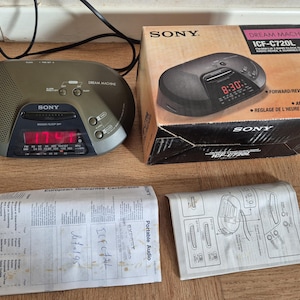 Może przedstawiać: Radiobudzik Sony Dream Machine ICF-C720L z ciemnoszarym, owalnym korpusem i czerwonym wyświetlaczem cyfrowym. Zestaw zawiera oryginalne kartonowe pudełko z tekstem, europejską kartę gwarancyjną oraz ilustrowaną instrukcję obsługi.