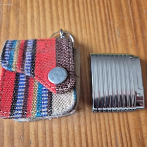 Könnte beinhalten: Ein kleines quadratisches Etui mit einem mehrfarbigen Streifenmuster in Rot, Blau und Beige, ausgestattet mit einem silbernen Metalldruckknopf und einem Schlüsselring, neben einem rechteckigen silbernen Feuerzeug mit vertikaler Riffelung.