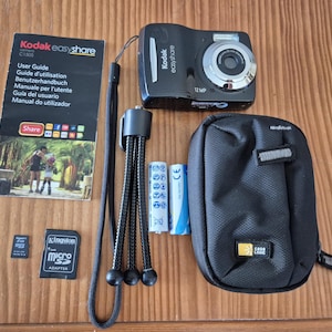 Op de afbeelding: Een zwarte Kodak EasyShare C1505 digitale camera met 12 MP lens, een gebruikershandleiding, een zwart flexibel statief, twee AA-batterijen, een microSD-kaart met adapter en een zwarte Case Logic tas met ritssluiting en een textuur van stof.
