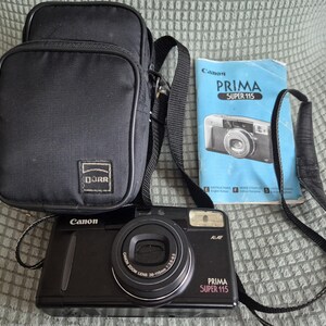 May include: A black Canon Prima Super 115 film camera with a zoom lens, a black carrying case with a Dürr logo, and a blue instruction manual. The camera features a built-in flash, a black neck strap, and text on the front reading Canon, Prima Super 115.