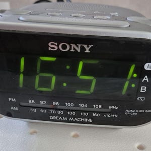 Może przedstawiać: Srebrne radiobudzik Sony Dream Machine z czarnym wyświetlaczem pokazującym godzinę 16:51 w zielonych cyfrach. Radio posiada opcje częstotliwości radiowych FM i AM oraz funkcję alarmu.