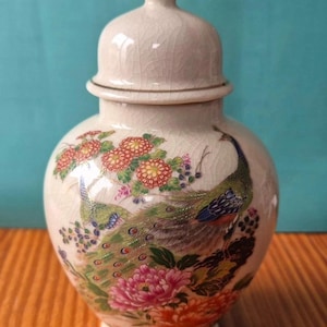 Peut inclure: Pot en céramique crème avec couvercle, orné d'un motif de paon. Le pot a un corps rond et un couvercle avec un bouton doré. Le paon est représenté avec des plumes vertes, bleues et dorées, entouré de fleurs.