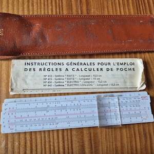 Peut inclure: Une règle à calcul vintage avec un étui en cuir et une notice. La règle à calcul est blanche avec des marquages noirs et rouges. L'étui en cuir est marron et porte le numéro "615". La notice est en français.